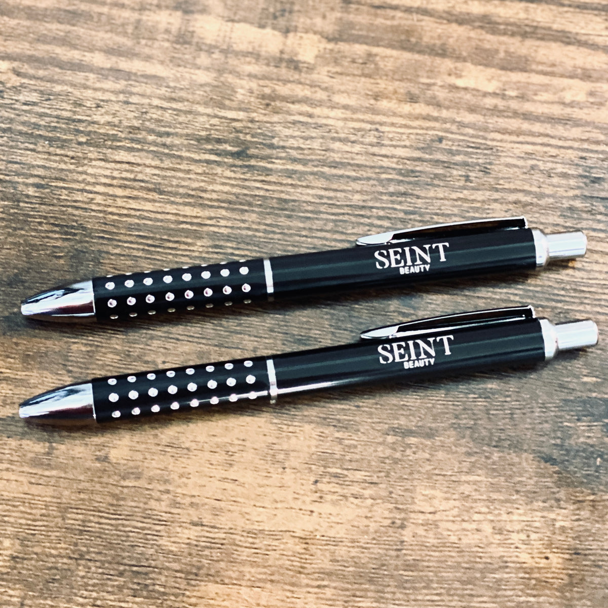 Two black Starlight pens engraved with Seint Beauty logo in silver, on rustic wood surface
